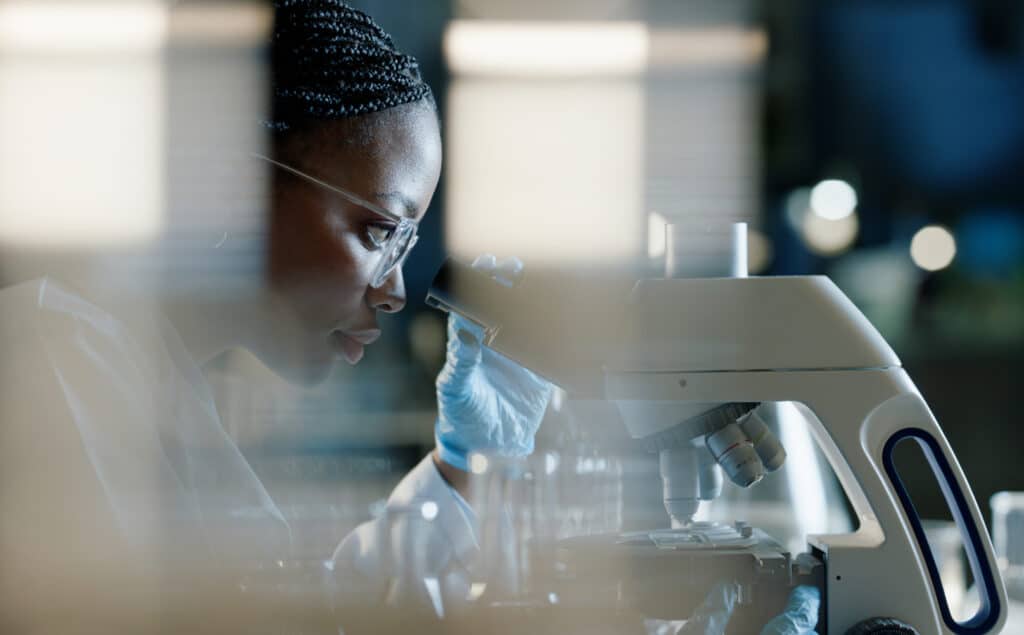A focused scientist in a lab coat examines samples through a microscope, surrounded by blurred laboratory equipment