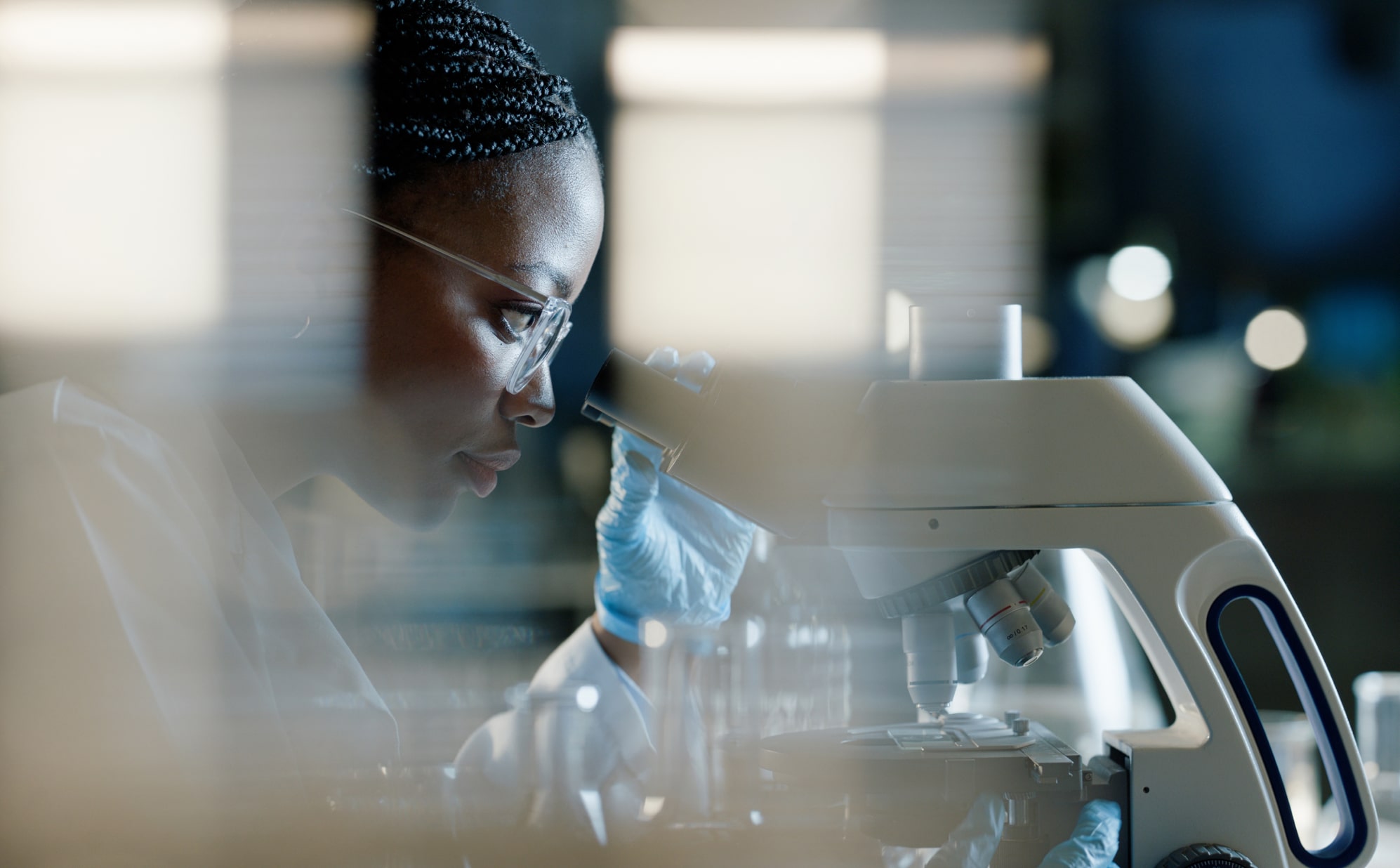 A focused scientist in a lab coat examines samples through a microscope, surrounded by blurred laboratory equipment