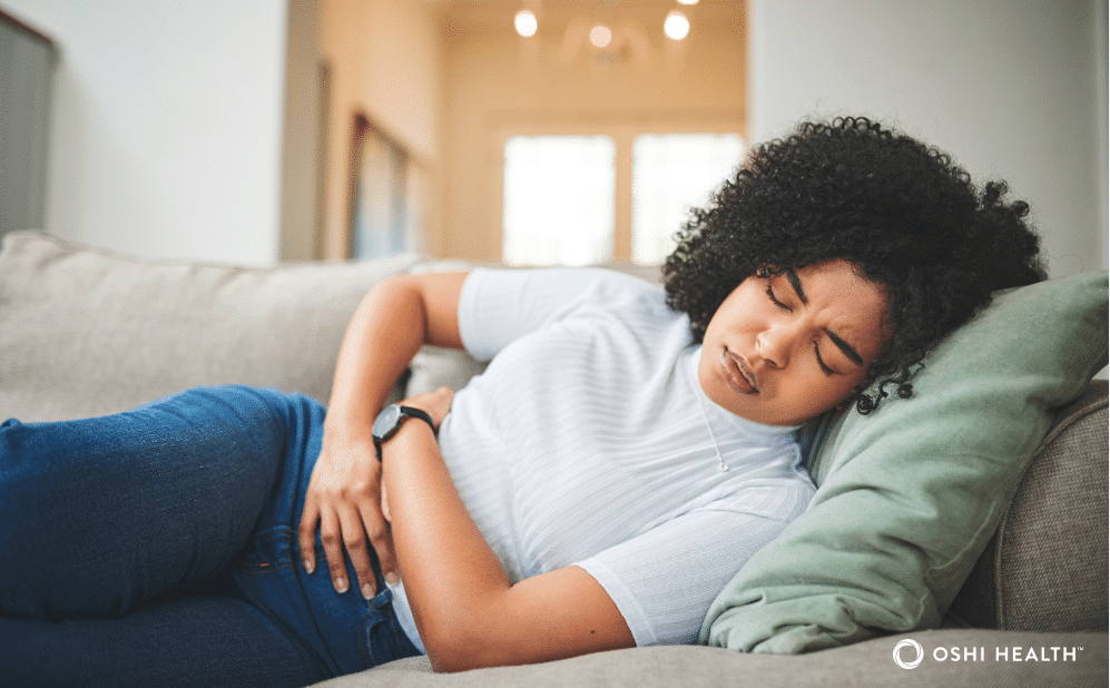 A woman lies on a couch holding her stomach and wincing in pain, suggesting abdominal discomfort.