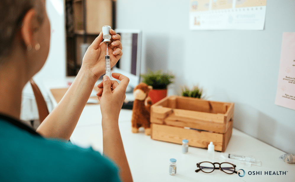 Person preparing an injection from a medication vial using a syringe at a desk, with additional medication supplies and glasses nearby.